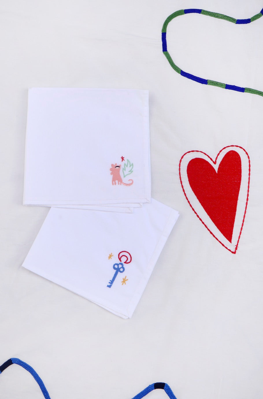 The first napkin has a pink lion with a green leaf and small red heart embroidered in the corner, and the other has a blue key with stars and a red moon outline. 