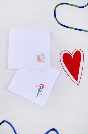 The first napkin has a pink lion with a green leaf and small red heart embroidered in the corner, and the other has a blue key with stars and a red moon outline. 