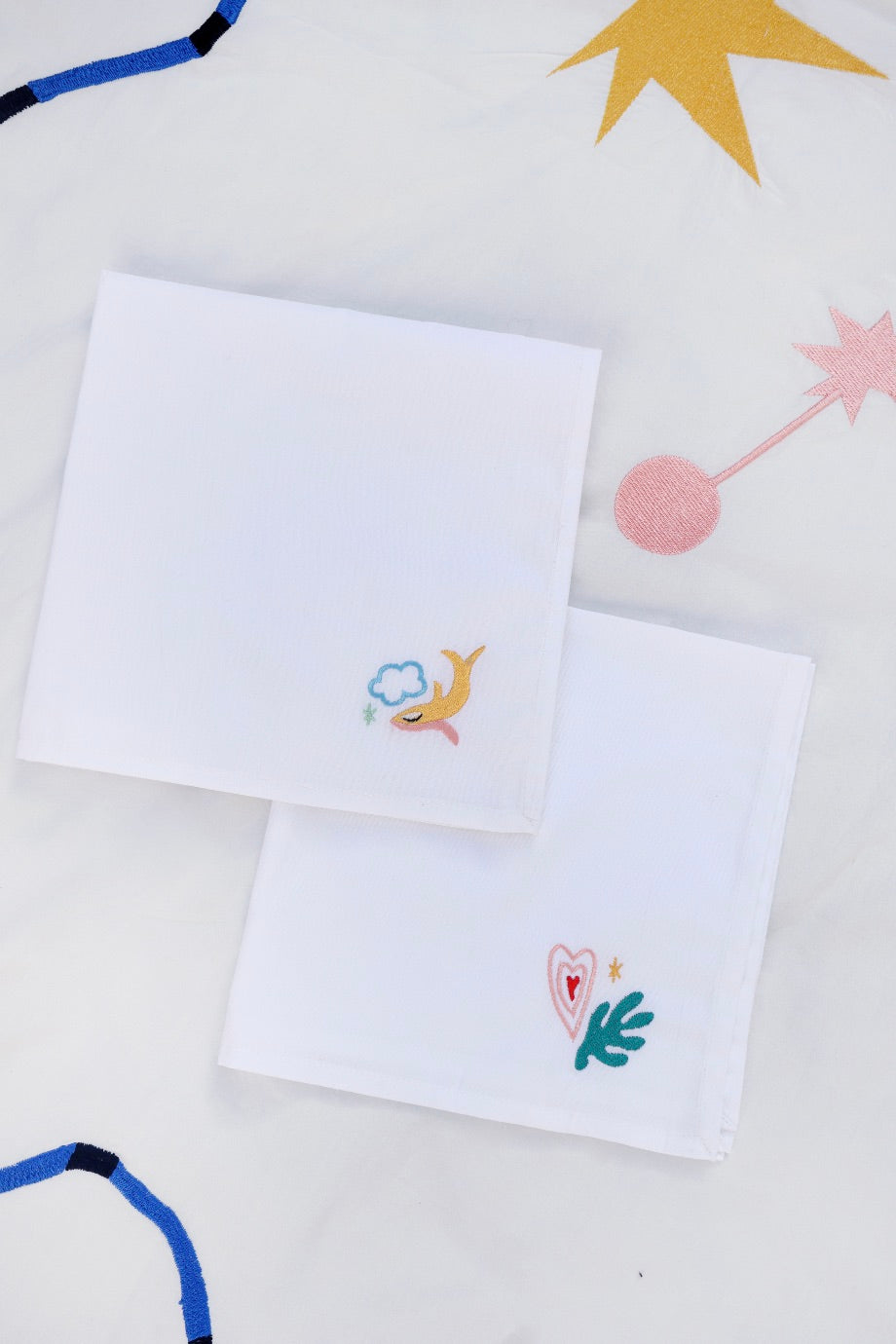 The first napkin has a yellow fish with the blue outline of a cloud, and the second has a pink and red heart and green filled in leaf embroidered.