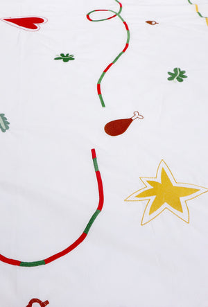 The white tablecloth has Christmas themed items embroidered on it: red heart, green leaves, green beans, carrots, chicken/turkey drumstick, a bright star and a red and green swirl covering the entire middle section.